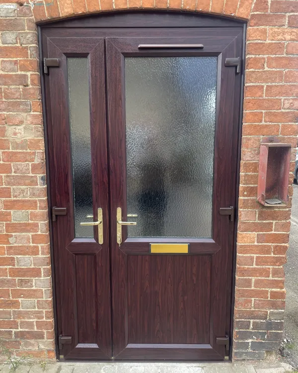 Rosewood-effect uPVC double doors with obscure glass and brass handles, fitted under a curved brick arch.