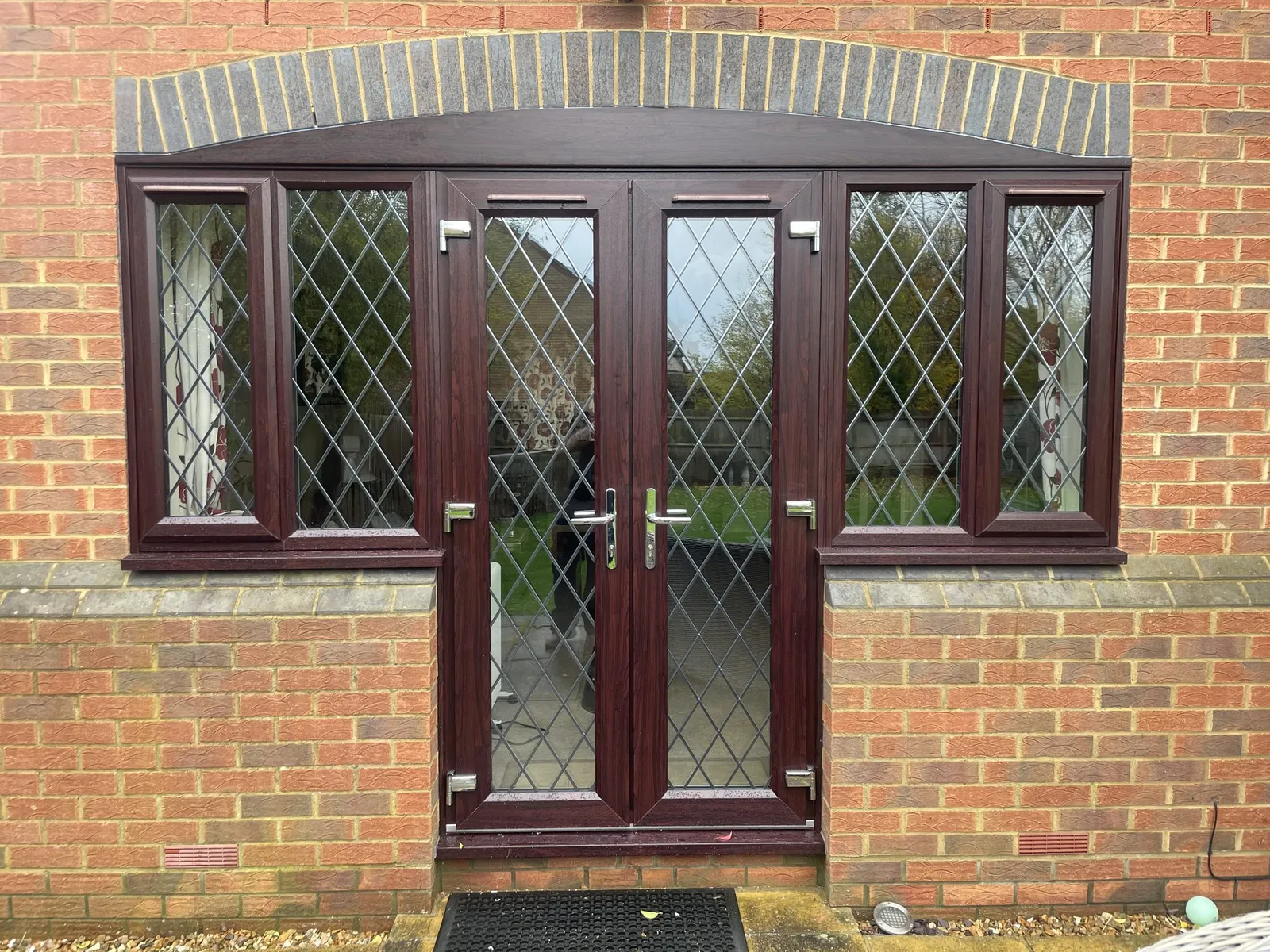 Rosewood-effect uPVC French doors with diamond leaded glass and matching leaded side windows under a brick arch.