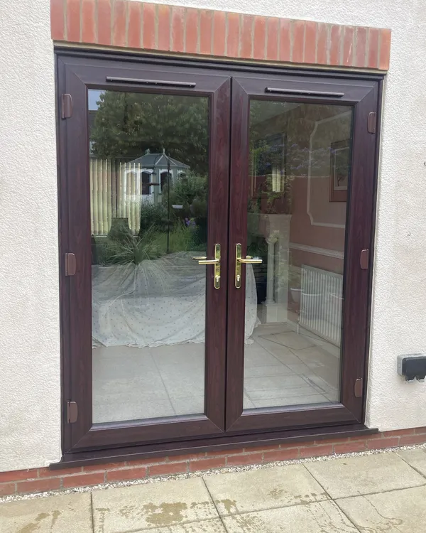 Rosewood-effect uPVC French doors with brass handles fitted in a white rendered wall under a brick header.