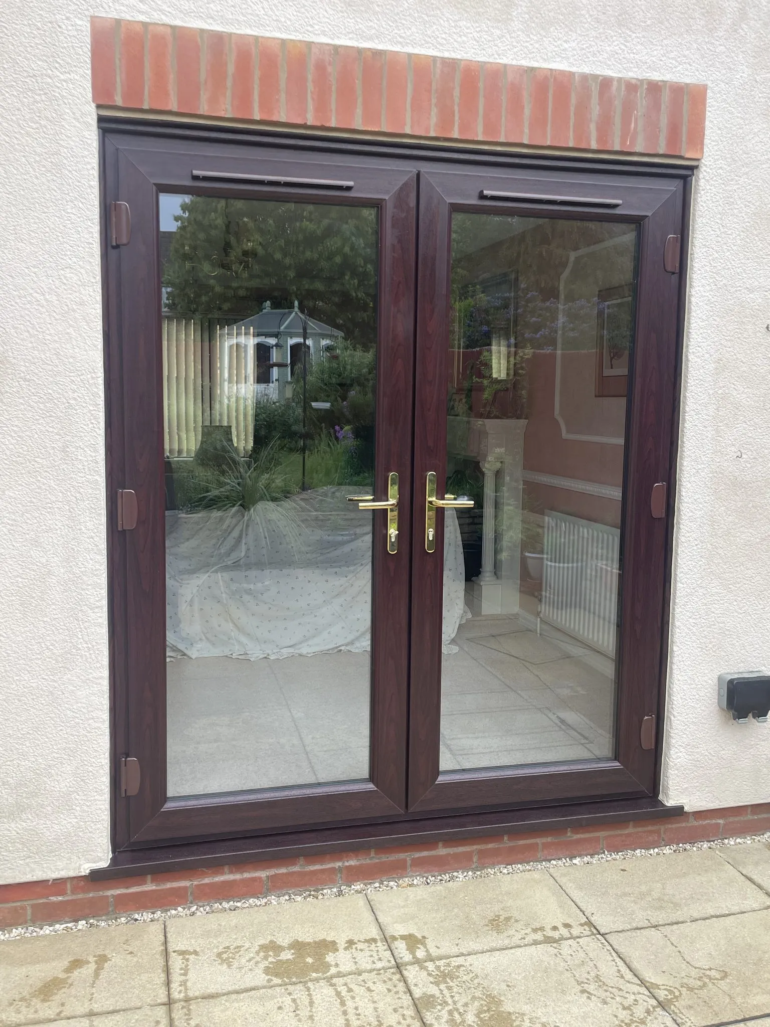 Rosewood-effect uPVC French doors with brass handles fitted in a white rendered wall under a brick header.