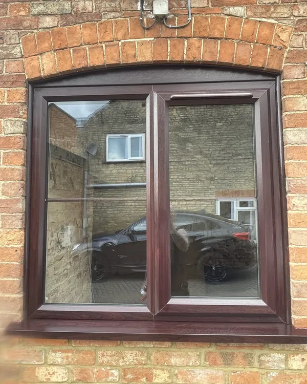Rosewood-effect uPVC casement window with a curved head, fitted beneath a brick arch in a red brick wall.