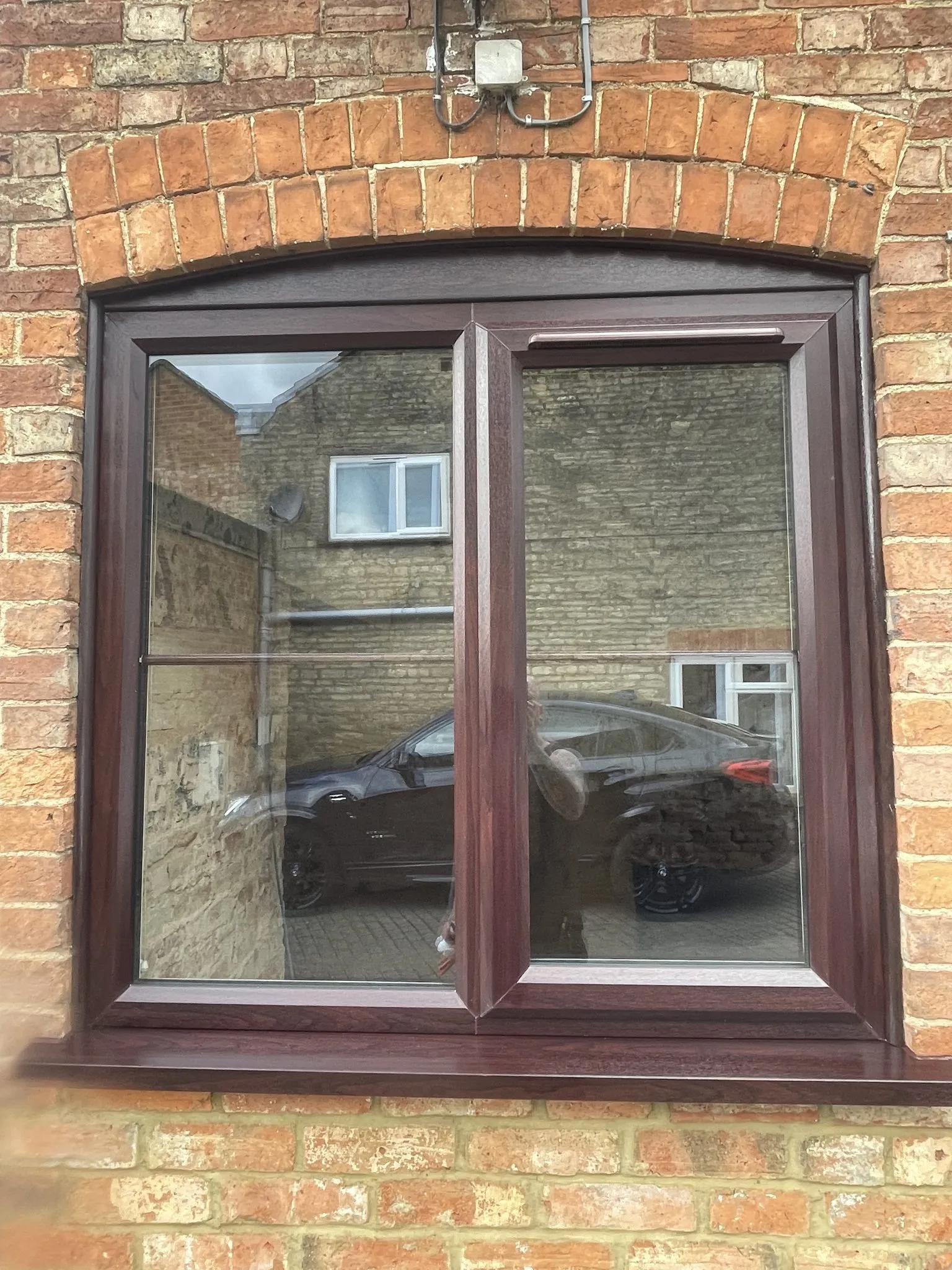 Rosewood-effect uPVC casement window with a curved head, fitted beneath a brick arch in a red brick wall.