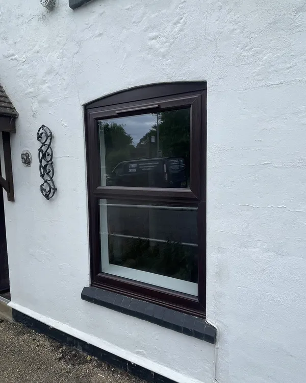 Rosewood-effect uPVC window with a curved head, fitted in the white rendered wall of a cottage.