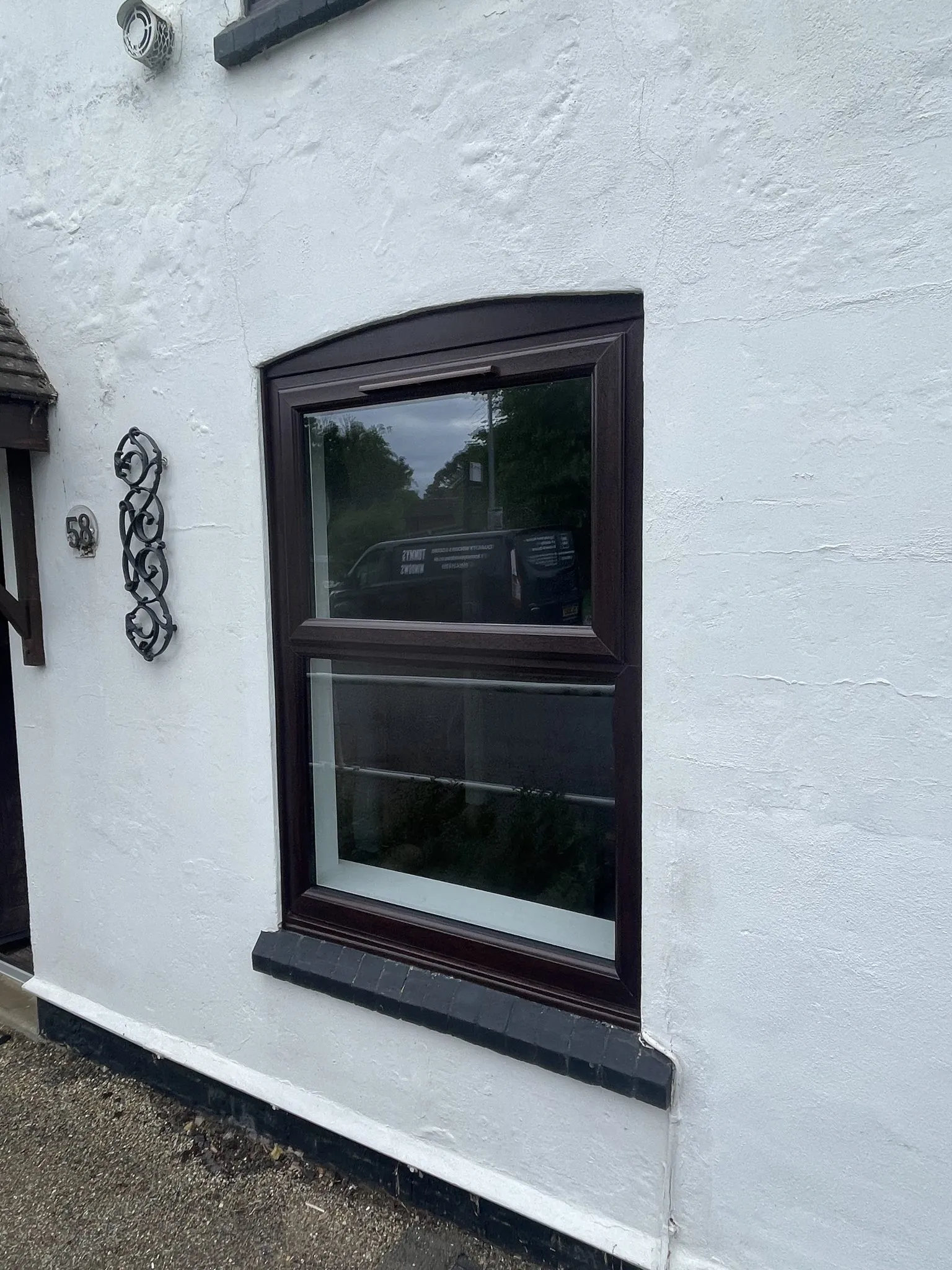 Rosewood-effect uPVC window with a curved head, fitted in the white rendered wall of a cottage.