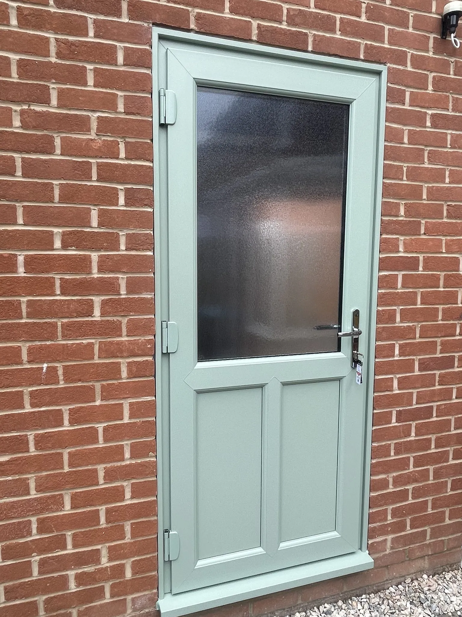 Sage green composite back door with an obscure glazed top panel and two moulded lower panels, in a red brick wall.