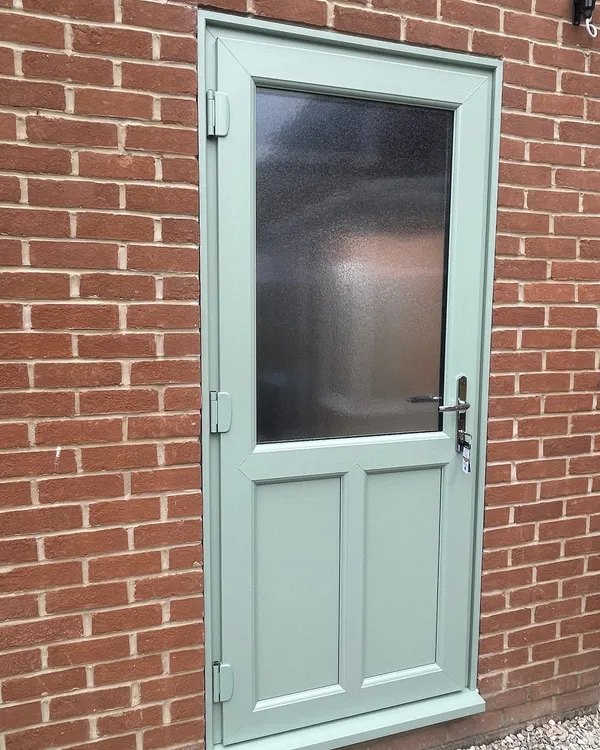 Sage green composite back door with an obscure glazed top panel and two moulded lower panels, in a red brick wall.