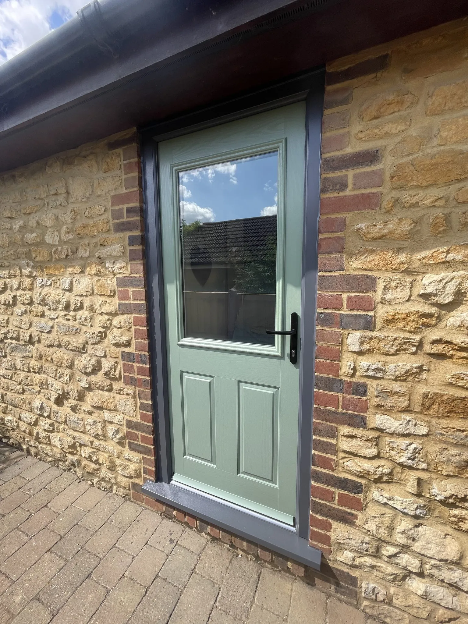 Sage green half-glazed composite door in an anthracite grey frame, set into an ironstone and brick wall.