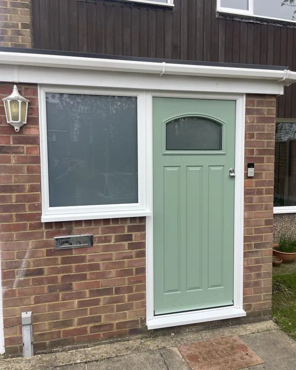 Sage green cottage-style composite door with a small arched glass panel beside a white uPVC window with obscure glass.