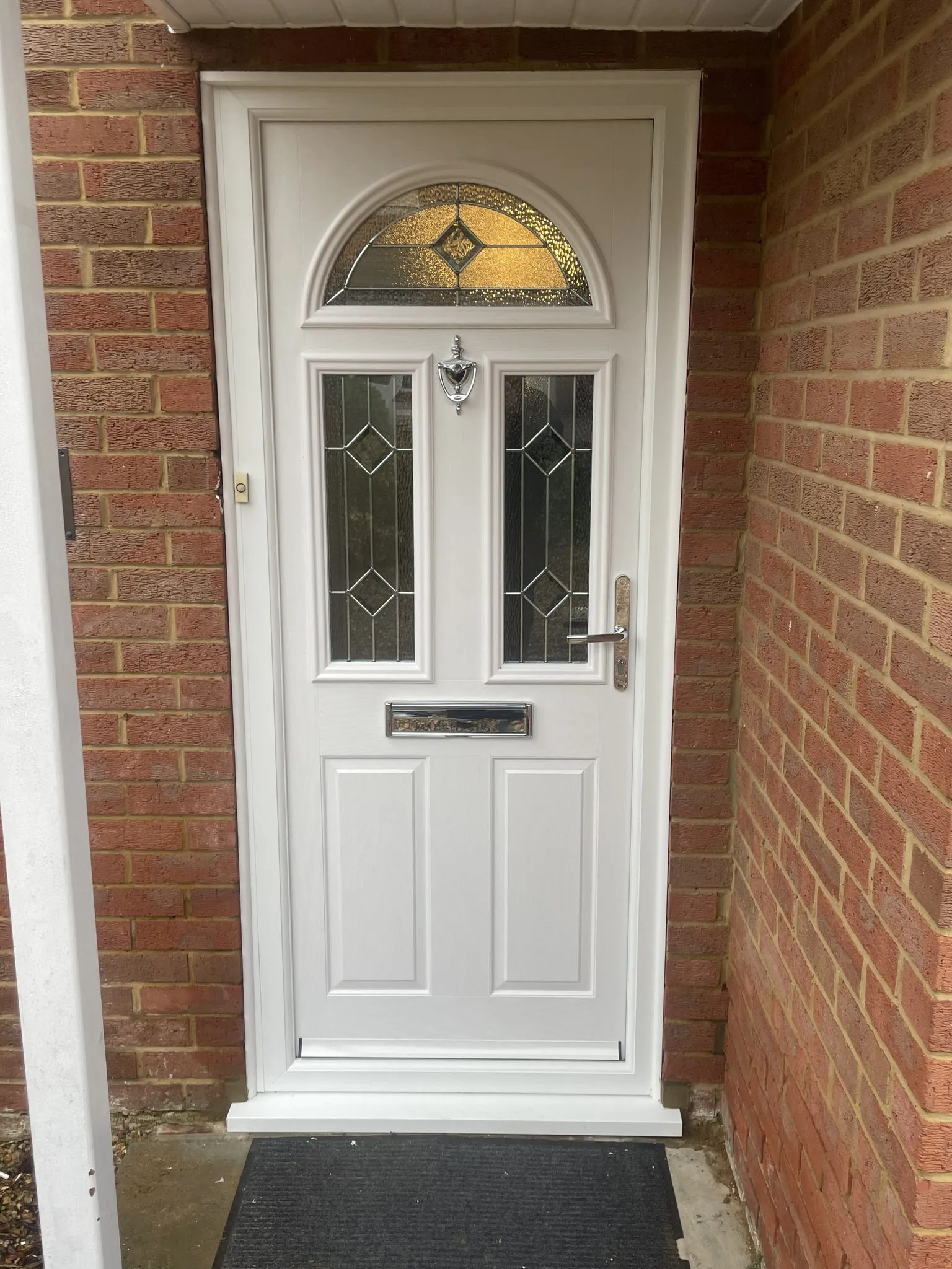 White composite front door with a half-moon leaded glass panel, two glazed panels and chrome furniture, inside a brick porch.