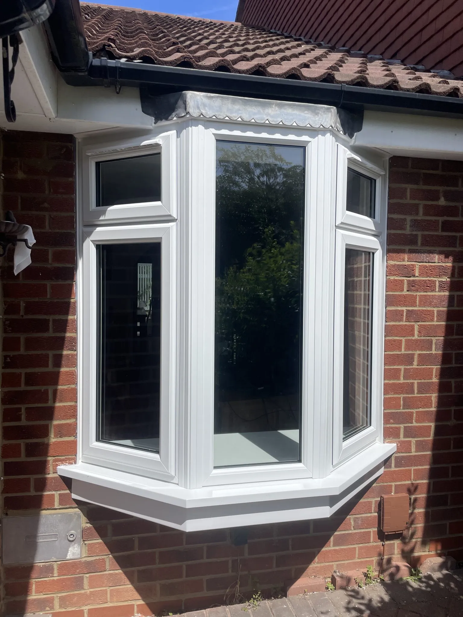 Three-sided white uPVC bay window with top openers fitted on a red brick house beneath a tiled roof.