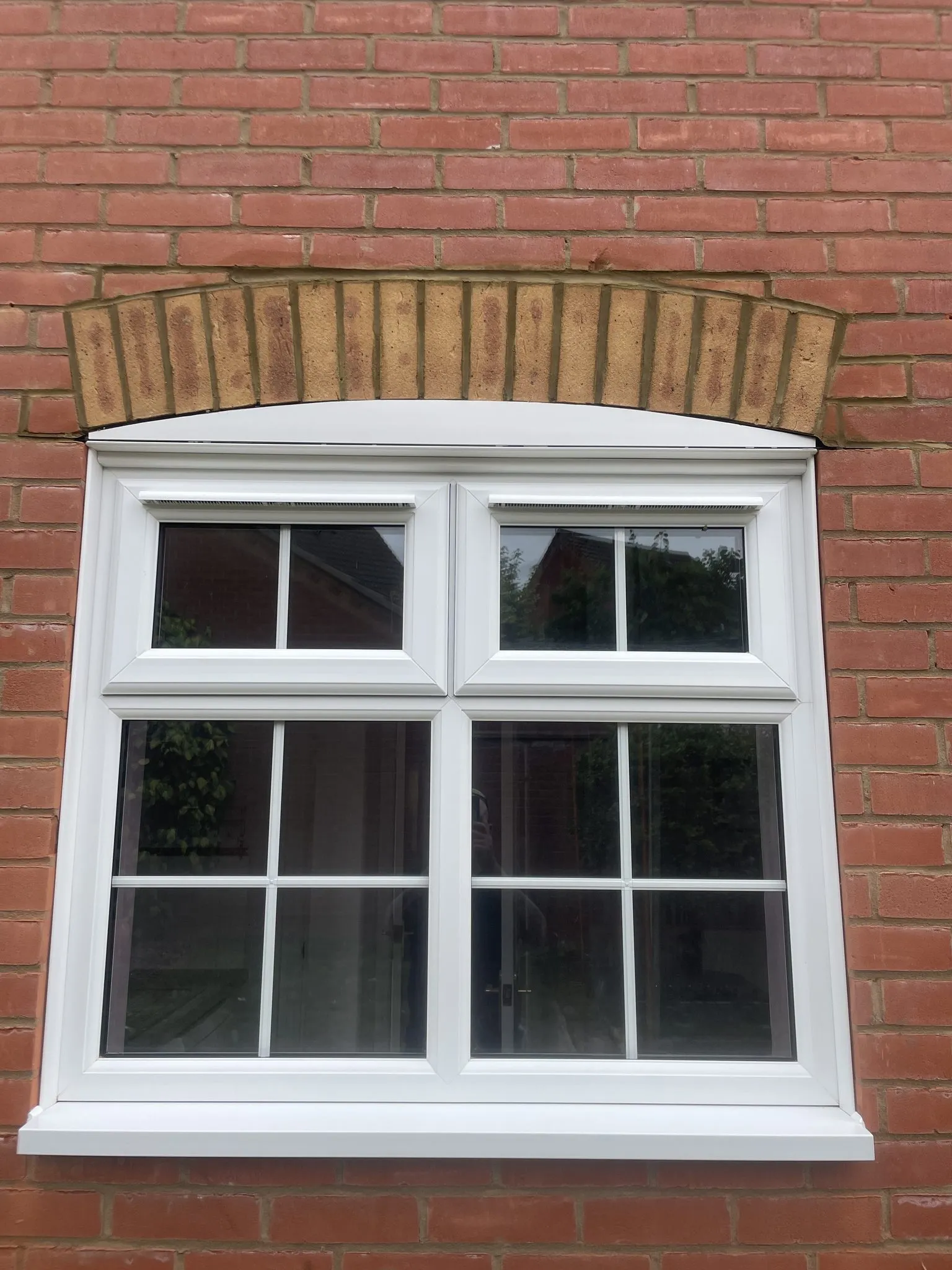 White uPVC double glazed casement window with Georgian bars and top openers, set beneath a curved brick arch in a red brick wall.