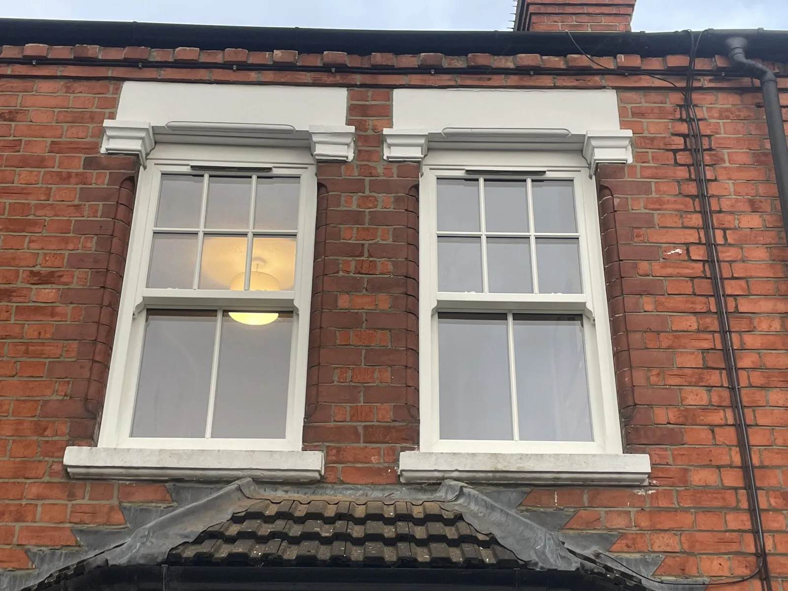 Two white uPVC sliding sash windows with Georgian bars and decorative heads above a bay roof on a Victorian red brick house.