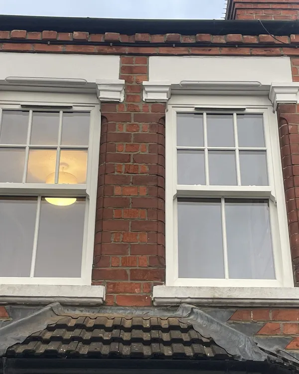 Two white uPVC sliding sash windows with Georgian bars and decorative heads above a bay roof on a Victorian red brick house.