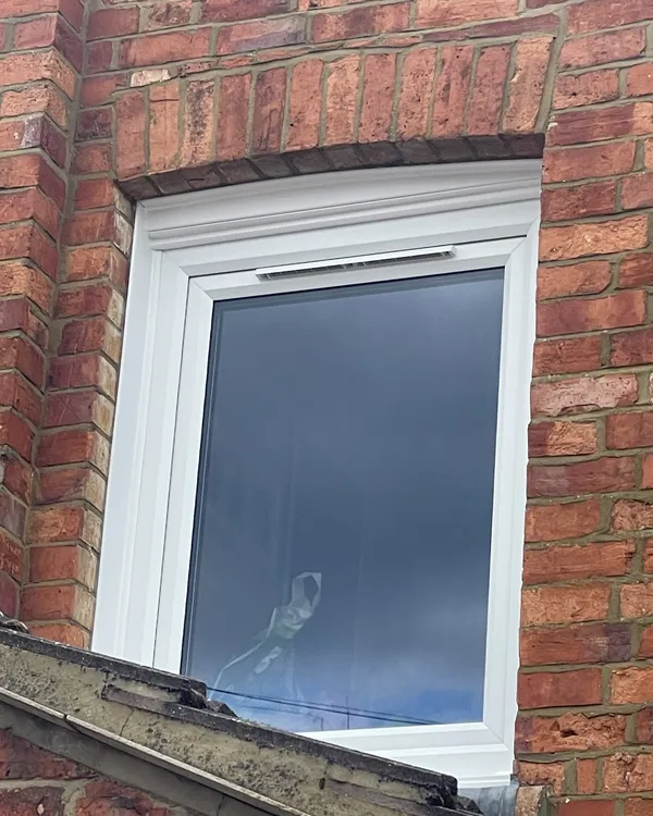 White uPVC window with a trickle vent fitted under a brick arch in an old red brick wall.
